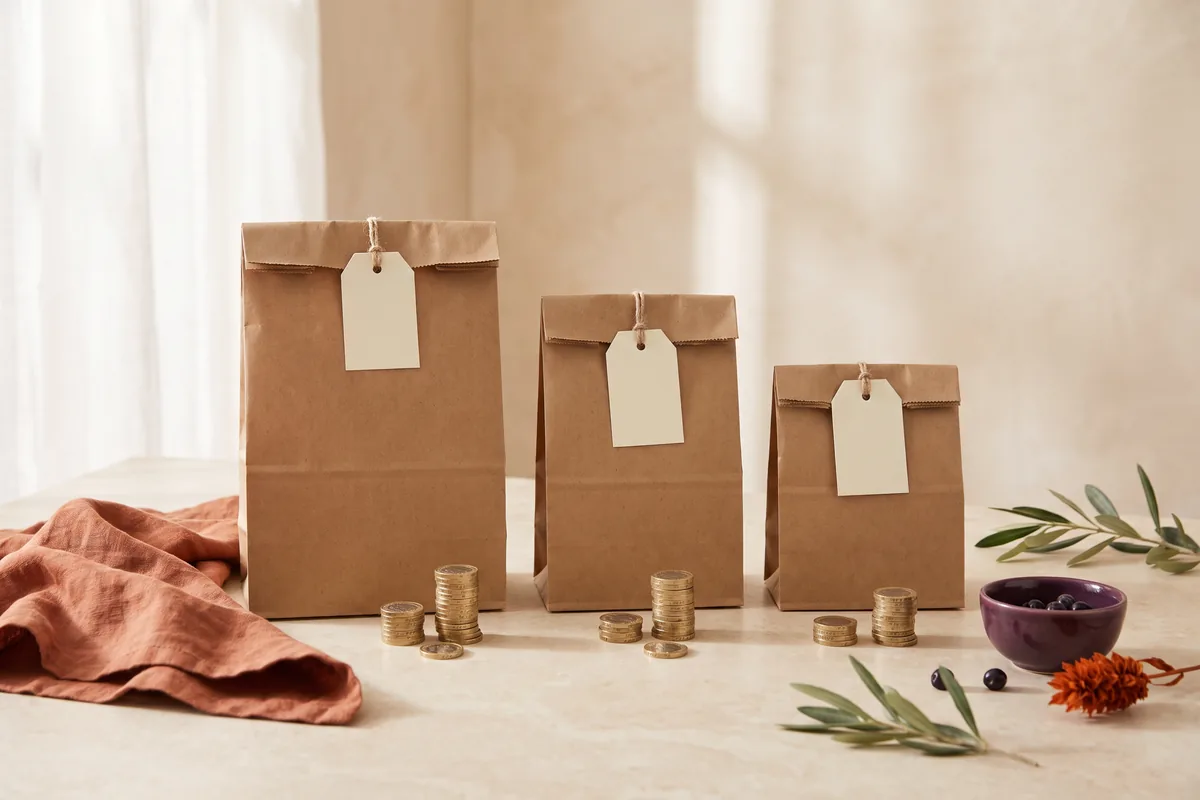 Three takeaway bags with blank tags, each with a different stack of pound coins in front.